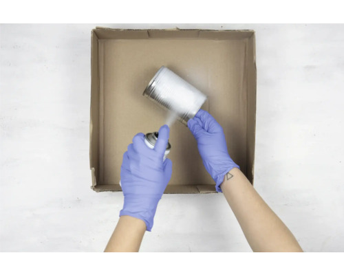 Une personne gantée vaporise de la peinture sur une boîte de conserve dans un carton.
