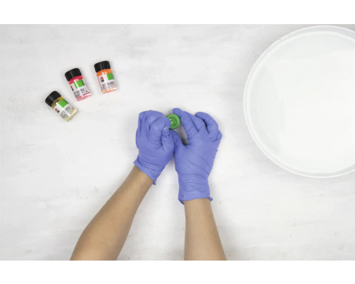 Une personne portant des gants ouvre un pot de peinture à marbrer, avec trois autres couleurs et un bol d''eau à côté.