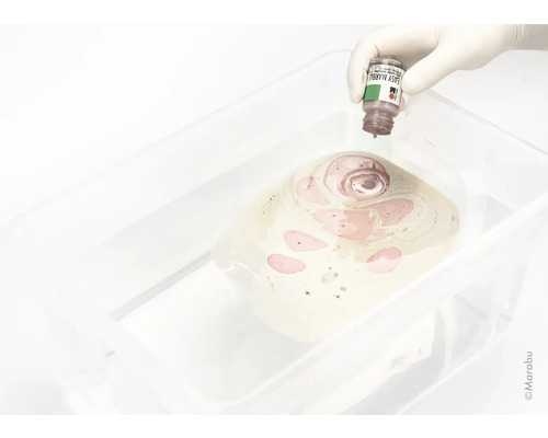 Eine Hand mit Handschuh träufelt Marmorierfarbe in ein Wasserbad für die Marmorierungstechnik.
