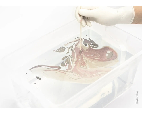 Eine Hand mit Handschuh marmoriert Farbe in einer Wanne mit einem Holzstab.