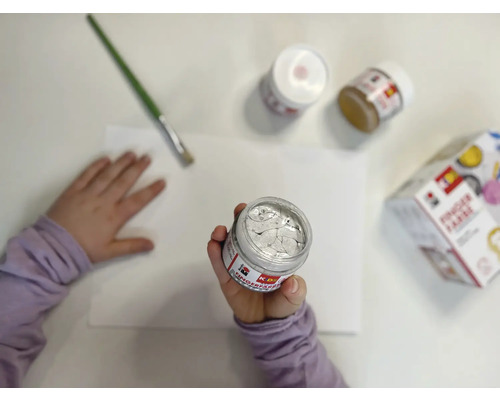 Kind hält Farbtopf über Papier, Pinsel und weitere Farbtöpfe liegen bereit