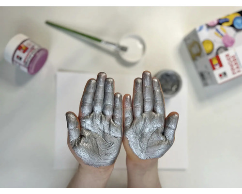 Zwei Hände mit silberner Farbe, Farbtiegel und Pinsel auf einem Tisch