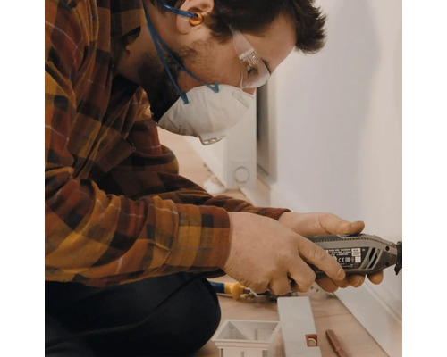 Un homme travaille avec un outil multifonctionnel sur une plinthe, protégé par un masque respiratoire et des lunettes de sécurité.