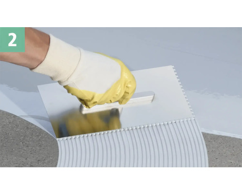 Eine Hand mit Schutzhandschuh verteilt Fliesenkleber mit einer Zahnspachtel.