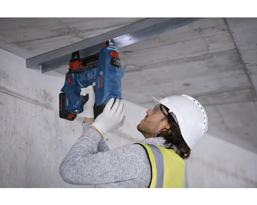 Un ouvrier fixe un rail métallique à un plafond en béton avec un cloueur à béton.