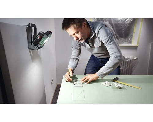 Un homme dessine au crayon des contours sur une surface verte, éclairée par une lampe de travail portative fixée au mur.