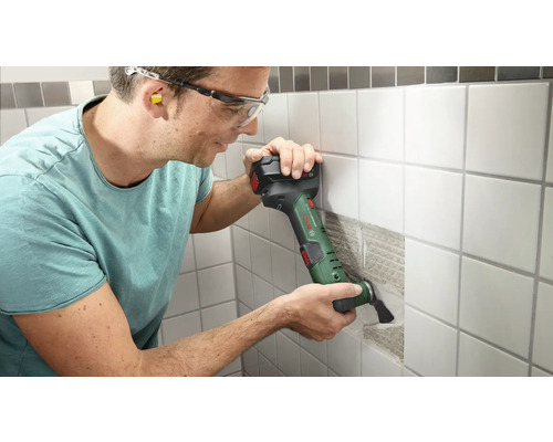 Un homme travaille avec un outil multifonctionnel sur un mur carrelé. Il porte des lunettes de protection et des bouchons d'oreille.