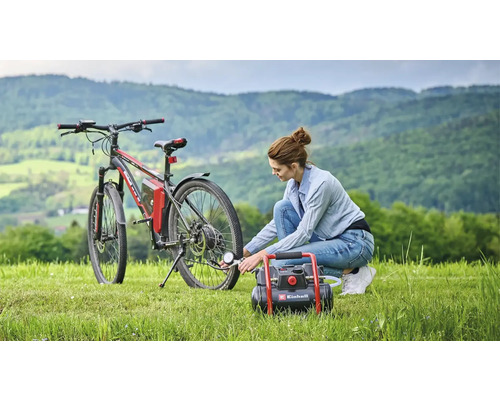 Une femme gonfle un pneu de vélo avec un compresseur Einhell dans une prairie devant un paysage de collines