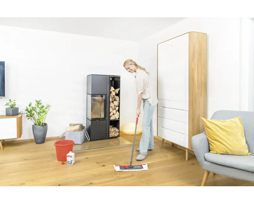 Femme nettoie le parquet avec un balai à franges dans un salon avec un poêle et des meubles.