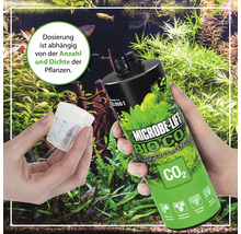 Microbe Lift Bio CO2 Flüssigdünger in einer Hand, Dosierbecher in der anderen, im Hintergrund ein Aquarium mit Pflanzen.