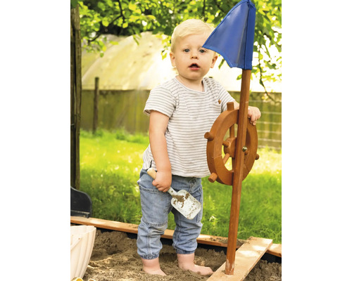 Kleinkind spielt mit einem Spielschiff mit Steuerrad und blauer Flagge im Sandkasten