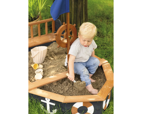 Kind spielt in einem Sandkasten in Schiffsform mit Ruder, Flagge und maritimen Motiven.