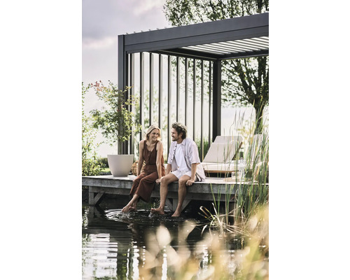 Un couple est assis sur un ponton en bois au bord de l''eau sous une pergola. Aménagement de jardin avec mobilier de salon et plantation.