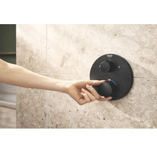 Une main actionne un thermostat de douche Grohe sur un mur carrelé