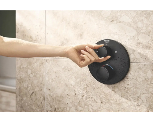 Main actionnant un thermostat de douche noir Grohe sur un mur carrelé