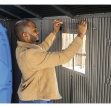 Ein Mann montiert ein Fenster in einem grauen Gartenhaus.