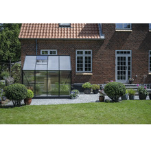 Gewächshaus aus Glas und Metall vor einem Backsteingebäude mit Ziegeldach und Rasenfläche.