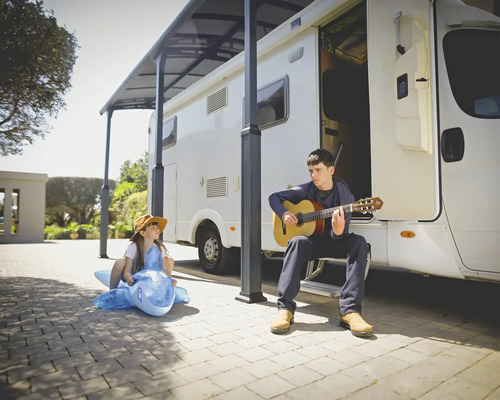 Un homme joue de la guitare devant un camping-car, tandis qu'un enfant est assis sur un dauphin gonflable.