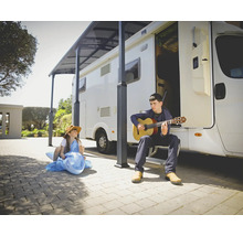 Un homme joue de la guitare devant un camping-car, tandis qu'un enfant est assis sur un dauphin gonflable.