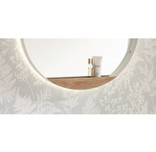 Miroir mural rond avec éclairage LED intégré et étagère incurvée en bois devant un mur au motif de fougères.