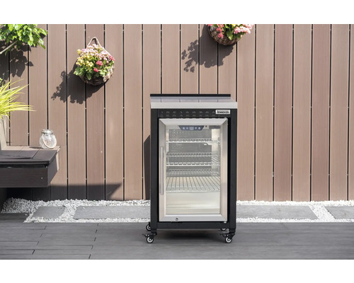 Réfrigérateur d extérieur Tenneker mobile avec porte vitrée et roulettes sur une terrasse devant une paroi en bois.