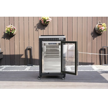 Réfrigérateur extérieur Tenneker noir avec porte vitrée ouverte et étagères métalliques sur une terrasse devant une clôture.
