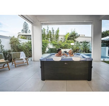 Jacuzzi carré moderne avec deux personnes sur une terrasse carrelée claire entourée de plantes de jardin.
