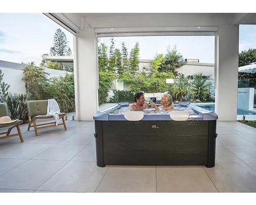 Jacuzzi carré moderne avec deux personnes sur une terrasse carrelée claire entourée de plantes de jardin.