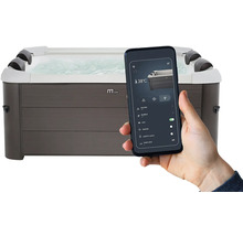 Main tenant un smartphone avec une application pour piloter un spa rectangulaire aspect bois gris, affichant des réglages tels que 38 degrés.