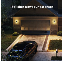 Wandleuchten mit Bewegungssensor an einer Garage bei Nacht, hell beleuchtete Einfahrt mit geparktem Auto und Text Täglicher Bewegungssensor.