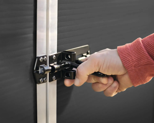 Main actionnant un verrou en métal noir sur un abri de jardin sombre avec un cadre argenté.