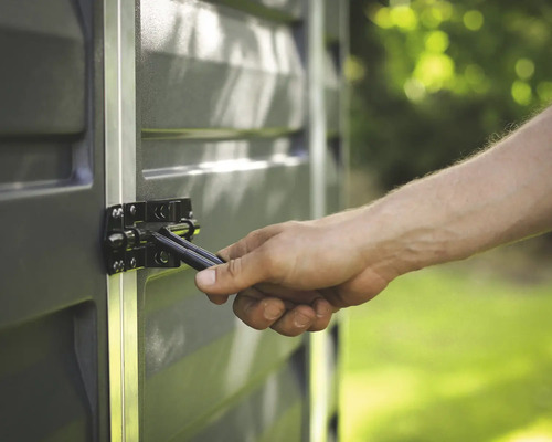 Gros plan d'une main actionnant un verrou métallique noir sur un abri de jardin gris avec des profilés en aluminium.