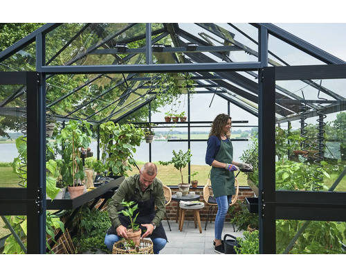 Geräumiges Gewächshaus aus Glas mit schwarzem Rahmen, zwei Personen pflegen Pflanzen in Töpfen, mit Blick auf einen See.