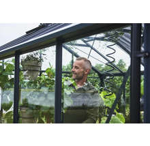 Ein Mann pflegt verschiedene Grünpflanzen in einem modernen Gewächshaus mit schwarzem Metallrahmen und großen Glasfenstern.