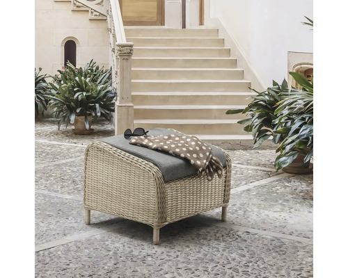 Geflochtener Gartenhocker mit grauem Kissen in einem hellen Innenhof mit Steintreppe und Pflanzen.