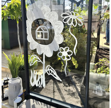 Fleurs stylisées et petite maison peintes en blanc sur la vitre d une serre, avec des plantes en arrière plan.