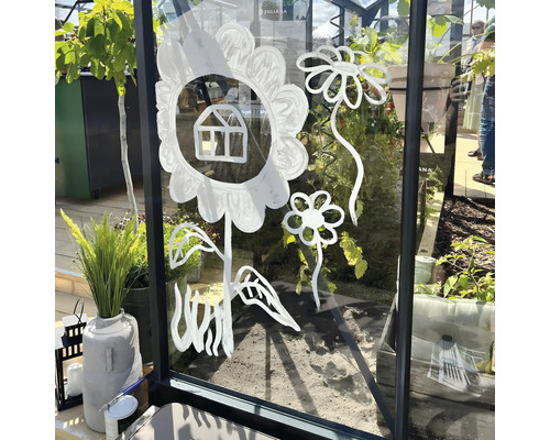 Fleurs stylisées et petite maison peintes en blanc sur la vitre d une serre, avec des plantes en arrière plan.