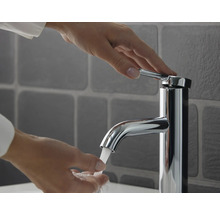 Eine Person bedient eine moderne verchromte Einhebel-Waschtischarmatur mit fließendem Wasser vor grauen Wandfliesen.