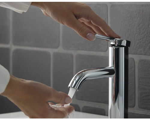 Eine Person bedient eine moderne verchromte Einhebel-Waschtischarmatur mit fließendem Wasser vor grauen Wandfliesen.