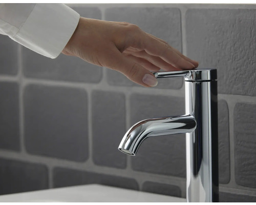 Eine Hand berührt eine verchromte Waschtischarmatur vor einer gefliesten Wand.