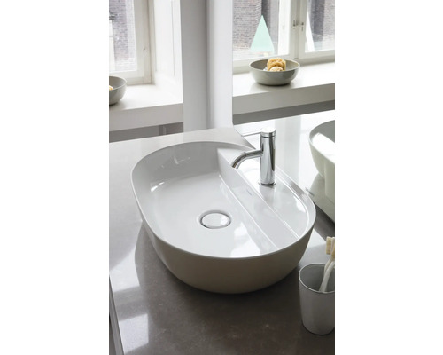 Ovales Aufsatzwaschbecken aus weißer Keramik mit verchromter Armatur in einem hellen, modernen Badezimmer auf einer Steinplatte.