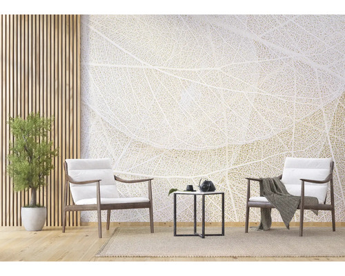 Salon lumineux avec papier peint panoramique motif nervures de feuilles, deux fauteuils en bois avec coussins blancs, table d'appoint noire et plante.