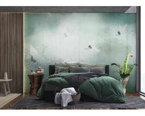 Papier peint photo en chambre avec grues blanches en forêt brumeuse, lit vert avec coussins, panneaux en bois au mur et plante verte.