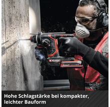 Une personne utilise un marteau perforateur sans fil Einhell sur un mur en béton avec des lunettes, un masque, un casque et des gants.