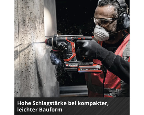 Une personne utilise un marteau perforateur sans fil Einhell sur un mur en béton avec des lunettes, un masque, un casque et des gants.