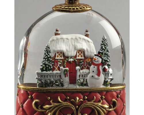 Schneekugel Weihnachtsdekoration mit verschneitem Haus, Schneemann und Tannen auf rotem Sockel mit goldenen Verzierungen.
