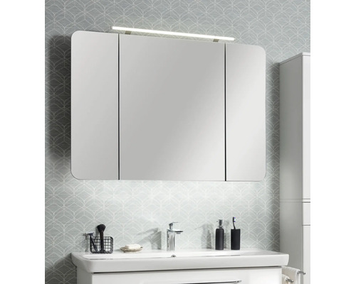 Armoire de toilette à trois portes avec coins arrondis et éclairage LED au-dessus d'un lavabo blanc dans une salle de bains lumineuse.