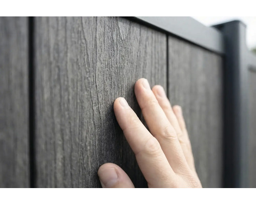 Une main touche la surface texturée d'un panneau de clôture aspect bois en composite bois-plastique.