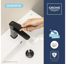 Main actionnant un mitigeur Grohe noir sur lavabo blanc. Symboles: QuickFix, outil, design allemand, cinq ans de garantie. Grohe Logo.