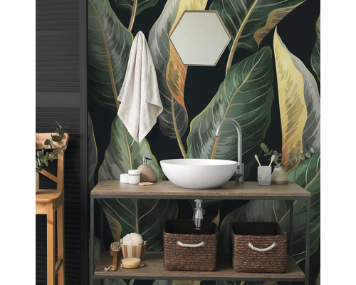 Papier peint photo avec de grandes feuilles botaniques dans une salle de bain derrière un meuble en bois avec vasque blanche et accessoires.